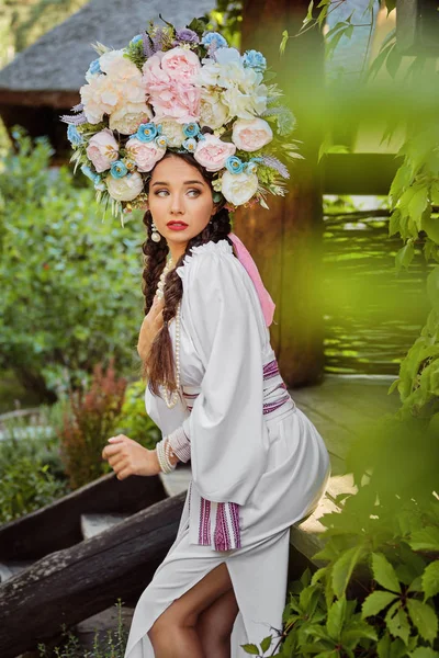 Bruna ragazza in un bianco ucraino autentico costume nazionale e una corona di fiori è in posa contro un cortile verde. Primo piano . — Foto Stock