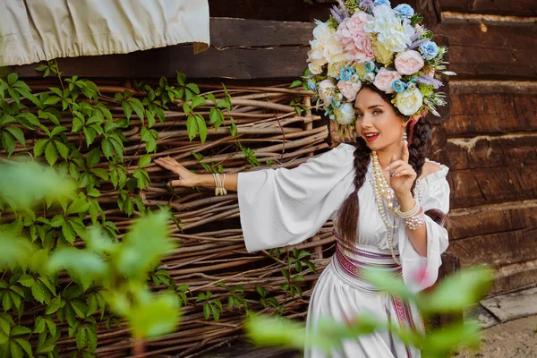 Barna lány egy fehér ukrán hiteles nemzeti ruha és a koszorú virágok pózol ellen terasz. — Stock Fotó