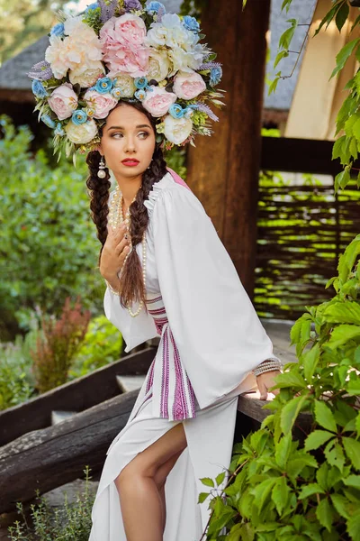 Ein brünettes Mädchen in weißem ukrainischen Nationalkostüm und einem Blumenkranz posiert vor einem grünen Hof. Nahaufnahme. — Stockfoto