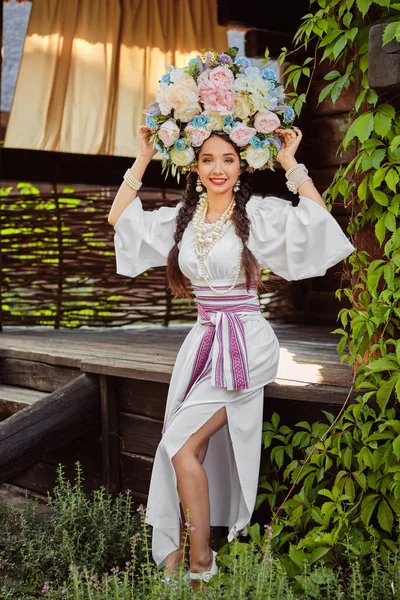 Brunette lány egy fehér ukrán hiteles nemzeti jelmez és a koszorú virág pózol ellen egy zöld udvar. — Stock Fotó