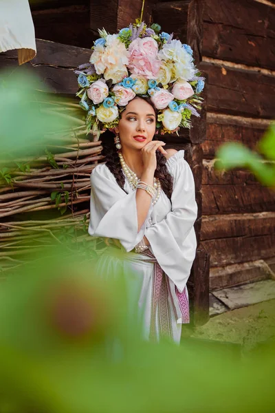 Bruna ragazza in un bianco ucraino autentico costume nazionale e una corona di fiori è in posa contro una terrazza. Primo piano . — Foto Stock