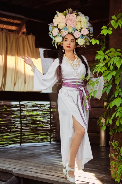 Chica morena en un traje nacional auténtico ucraniano blanco y una corona de flores está posando contra un patio verde . —  Fotos de Stock