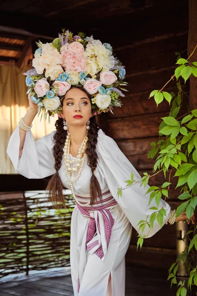 Brunetka dziewczyna w białym ukraińskim autentycznym Narodowym stroju i Wieniec kwiatów stwarza przeciwko zielonym podwórku. Zbliżenie. — Zdjęcie stockowe