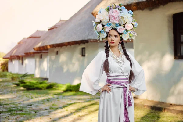 Chica morena en un traje nacional auténtico ucraniano blanco y una corona de flores está posando contra una choza blanca . —  Fotos de Stock