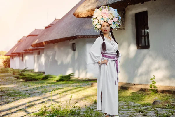 Chica morena en un traje nacional auténtico ucraniano blanco y una corona de flores está posando contra una choza blanca . —  Fotos de Stock