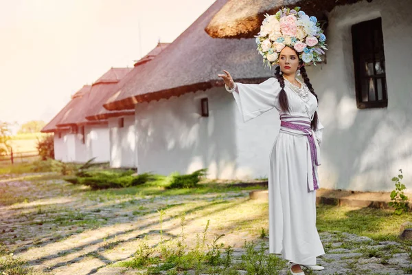Brunette lány egy fehér ukrán hiteles nemzeti jelmez és a koszorú virág pózol ellen fehér kunyhó. — Stock Fotó