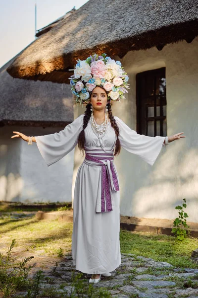 Дівчина брюнетка в білому українському автентичному національному костюмі і вінок з квітів позує проти білої хатини. — стокове фото