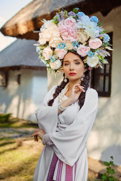 Bruna ragazza in un bianco ucraino autentico costume nazionale e una corona di fiori è in posa contro una capanna bianca. Primo piano . — Foto Stock