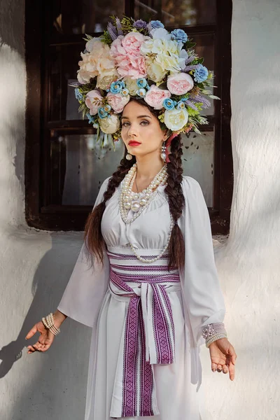 Дівчина брюнетка в білому українському автентичному національному костюмі і вінок з квітів позує проти білої хатини. Закри. — стокове фото