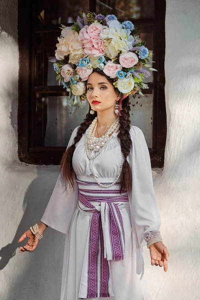 Bruna ragazza in un bianco ucraino autentico costume nazionale e una corona di fiori è in posa contro una capanna bianca. Primo piano . — Foto Stock