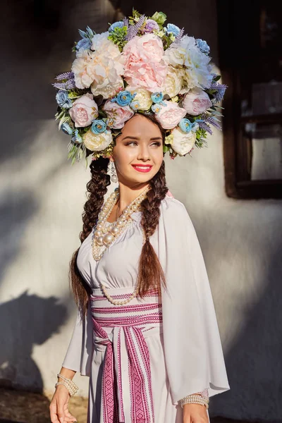 Дівчина брюнетка в білому українському автентичному національному костюмі і вінок з квітів позує проти білої хатини. Закри. — стокове фото