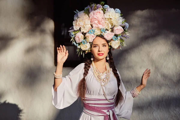 Bruna ragazza in un bianco ucraino autentico costume nazionale e una corona di fiori è in posa contro una capanna bianca. Primo piano . — Foto Stock