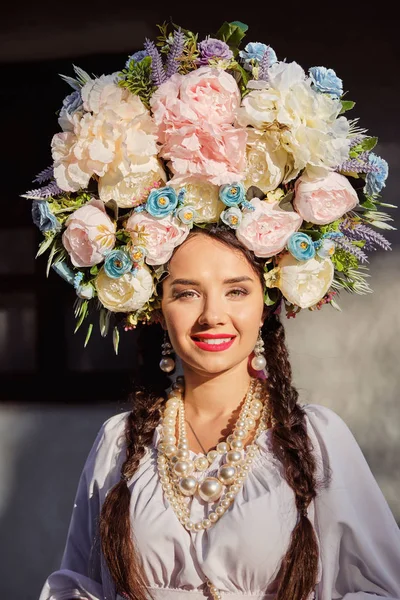 Brunette girl in a white ukrainian authentic national costume and a wreath of flowers is posing against a white hut. Close-up. — Stock Photo, Image