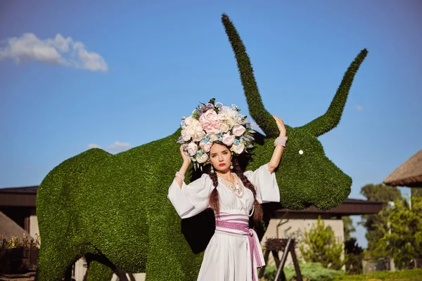 Brünettes Mädchen in weißem ukrainischen Nationalkostüm und einem Blumenkranz posiert vor einem grünen Ochsen, der aus einem Busch geschnitzt wurde. — Stockfoto