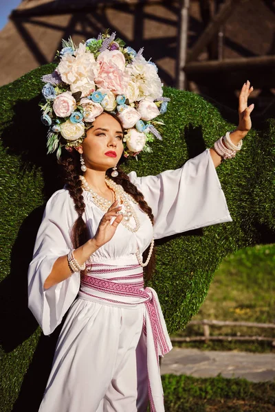 Brunetka dziewczyna w białym ukraińskim autentycznym Narodowym stroju i Wieniec kwiatów stwarza przeciwko zielony wół rzeźbione z krzewu. — Zdjęcie stockowe