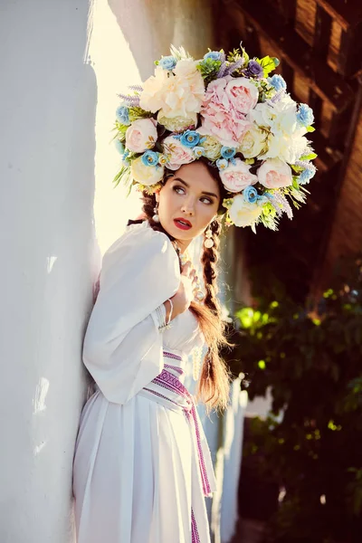 Brunette girl in a white ukrainian authentic national costume and a wreath of flowers is posing against a white hut. — Stock Photo, Image