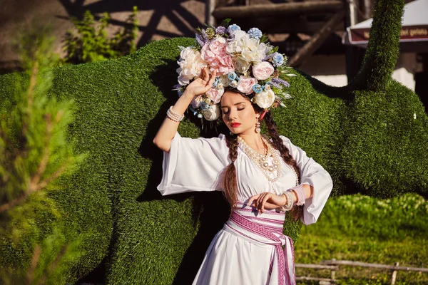 Barna lány egy fehér ukrán hiteles nemzeti ruha és a koszorút a virágok pózol ellen zöld ökör faragott egy bokor. — Stock Fotó