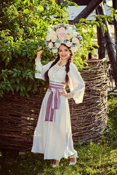 Bruna ragazza in un bianco ucraino autentico costume nazionale e una corona di fiori è in posa contro un recinto di vimini in legno . — Foto Stock