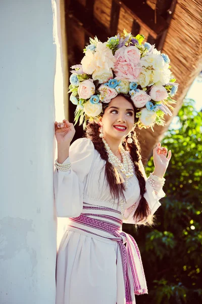 Brunette girl in a white ukrainian authentic national costume and a wreath of flowers is posing against a white hut. — Stock Photo, Image