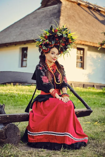 Brunett tjej i en vit ukrainsk autentisk nationell kostym och en krans av blommor poserar sittande på en träbänk mot en vit koja. — Stockfoto
