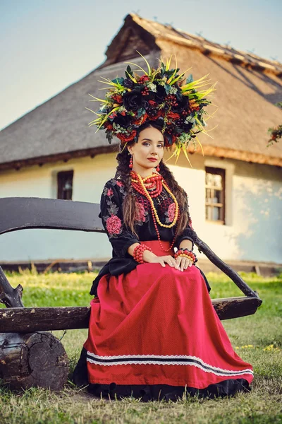 Brunett tjej i en vit ukrainsk autentisk nationell kostym och en krans av blommor poserar sittande på en träbänk mot en vit koja. — Stockfoto