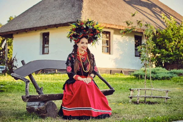 Brunette girl in a white ukrainian authentic national costume and a wreath of flowers is posing sitting on a wooden bench against a white hut. — Stock Photo, Image