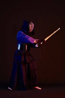 Yakın çekim, Kendo dövüşçüsü zırh giyiyor, geleneksel kimono giyiyor, miğfer takıyor shinai bambu kılıcıyla dövüş sanatları yapıyor, siyah arka plan.