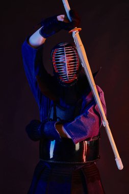 Yakın çekim, Kendo dövüşçüsü zırh giyiyor, geleneksel kimono giyiyor, miğfer takıyor shinai bambu kılıcıyla dövüş sanatları yapıyor, siyah arka plan.