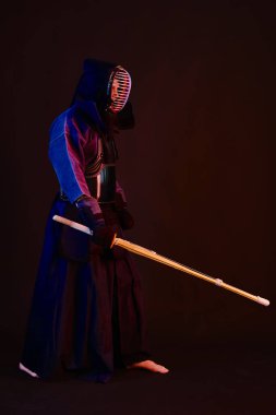 Yakın çekim, Kendo dövüşçüsü zırh giyiyor, geleneksel kimono giyiyor, miğfer takıyor shinai bambu kılıcıyla dövüş sanatları yapıyor, siyah arka plan.
