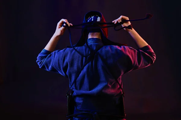 Feche o tiro, Kendo lutador vestindo em uma armadura e quimono tradicional está amarrando o lacado em seu capacete de volta contra um fundo preto. — Fotografia de Stock