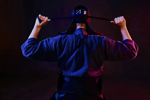 Cerrado el tiro, el luchador de Kendo con armadura y kimono tradicional está atando la laca de su casco de pie contra un fondo negro.. —  Fotos de Stock