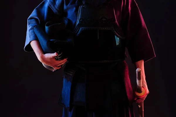 Kendo luchador usando una armadura, el kimono tradicional mantiene su casco y espada de bambú shinai mientras se pone sobre un fondo negro. Cierre. —  Fotos de Stock