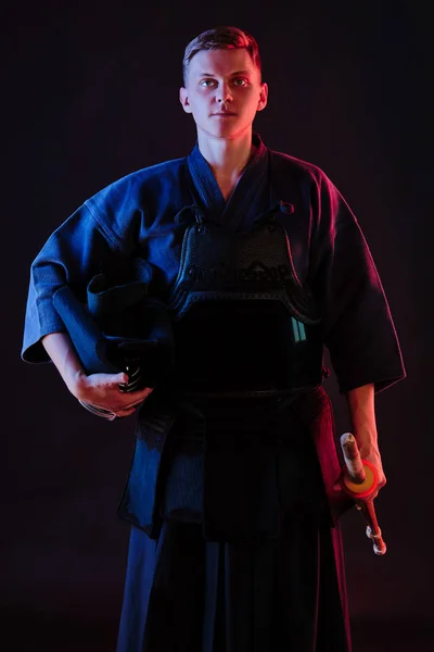 Kendo vechter draagt in een harnas, traditionele kimono houdt zijn helm en shinai bamboe zwaard terwijl hij poseert op een zwarte achtergrond. Sluiten.. — Stockfoto