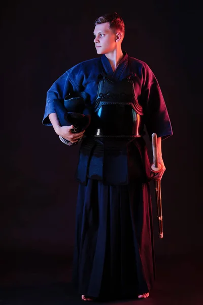 Боец кендо в доспехах, традиционном кимоно держит в руках шлем и меч синай-бу, позируя на черном фоне. Закрыть. — стоковое фото