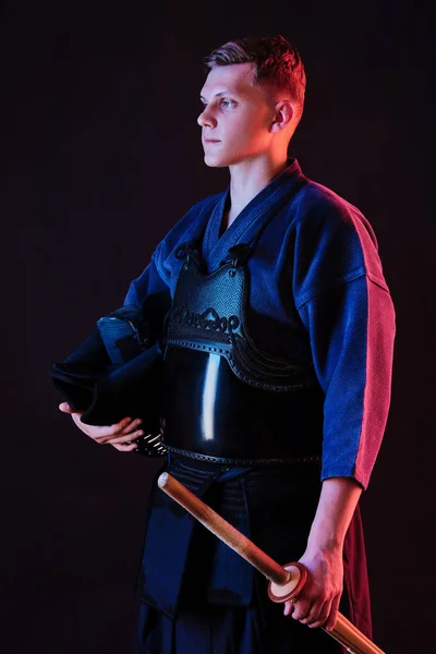 Kendo fighter wearing in an armor, traditional kimono is holding his helmet and shinai bamboo sword while posing on a black background. Close up. Stock Picture