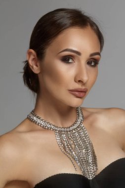 Siyah sutyen ve kolye takıp, gri arka planda poz veren esmer model bir kızın yakın plan portresi..