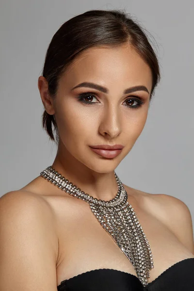 Close up portrait of a brunette model girl with professional evening make-up, wearing a black bra and necklace, posing on gray background.