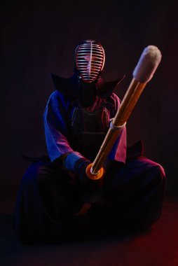 Kapatın. Kendo dövüşçüsü zırh giyiyor, geleneksel kimono giyiyor, miğfer takıyor, oturuyor, Shinai bambu kılıcıyla dövüş sanatları yapıyor, siyah arka planda..