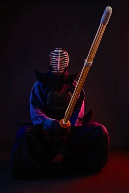 Kapatın. Kendo dövüşçüsü zırh giyiyor, geleneksel kimono giyiyor, miğfer takıyor, oturuyor, Shinai bambu kılıcıyla dövüş sanatları yapıyor, siyah arka planda..