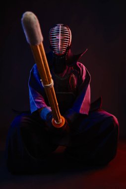 Kapatın. Kendo dövüşçüsü zırh giyiyor, geleneksel kimono giyiyor, miğfer takıyor, oturuyor, Shinai bambu kılıcıyla dövüş sanatları yapıyor, siyah arka planda..