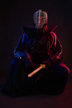 Kapatın. Kendo dövüşçüsü zırh giyiyor, geleneksel kimono giyiyor, miğfer takıyor, oturuyor, Shinai bambu kılıcıyla dövüş sanatları yapıyor, siyah arka planda..