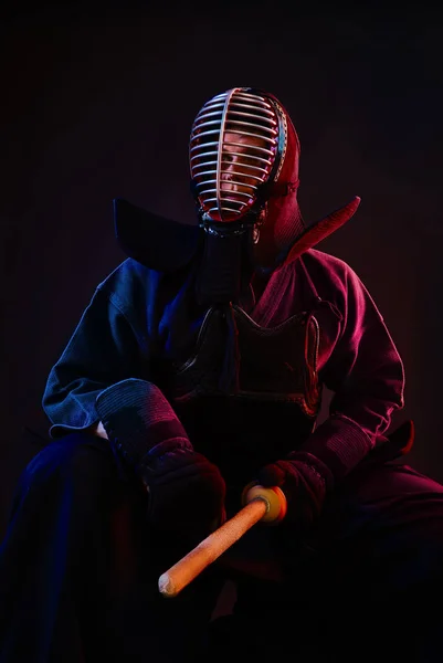 Close up. Kendo fighter wearing in an armor, traditional kimono, helmet, sitting, practicing martial art with shinai bamboo sword, black background. — Stock Photo, Image