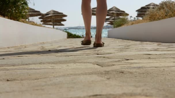 Em movimento, vista para trás de uma mulher loira em suite e túnica caminhando para a praia através de um caminho ao longo do mar mediterrâneo azul, Chipre . — Vídeo de Stock