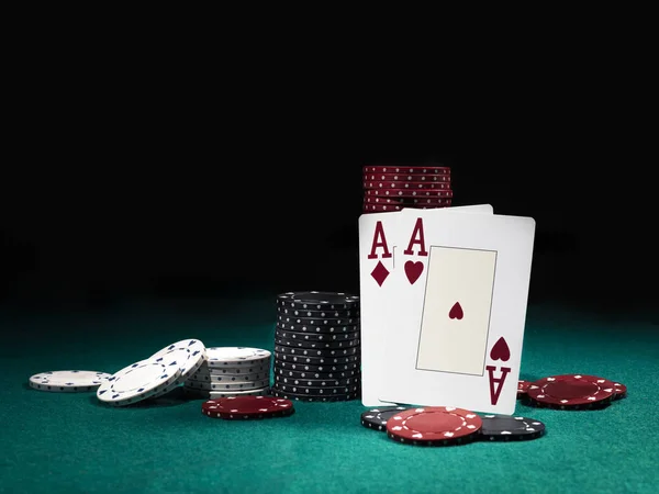 Two aces hearts and diamonds standing leaning on chips piles, some of them laying nearby on green cover of playing table. Black background. Close-up. — Stock Photo, Image