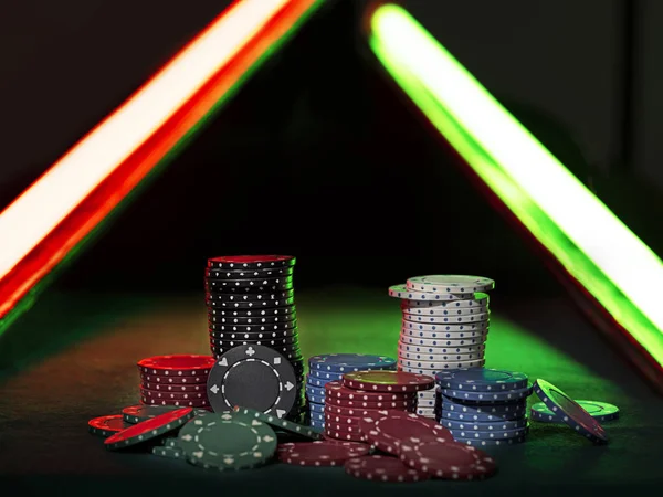 Close-up photo of a multicolored chips piles, some of them laying nearby on green cover of playing table, under green and red neon lights. Black background. Close-up. — Stock Photo, Image