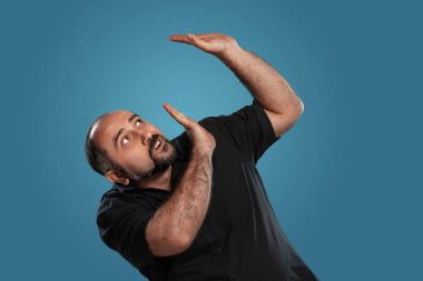 Siyah tişört giymiş sakallı, esmer, olgun bir erkeğin yakın plan portresi. Mavi bir stüdyo geçmişine karşı poz verirken üzerine bir şey düşeceğinden korkuyor. Samimi duygular konsepti. Boşluğu kopyala.