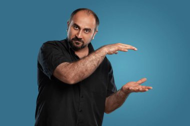 Siyah tişört giymiş, esmer ve sakallı bir yetişkinin yakın plan portresi. Kameraya bakıyor ve mavi bir stüdyonun arka planında poz verirken el kol hareketi yapıyor. Samimi duygular konsepti. Boşluğu kopyala.