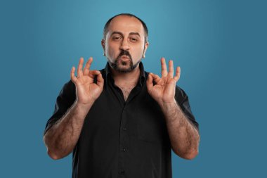 Siyah tişört giymiş esmer, sakallı, yakışıklı bir adamın yakın plan portresi. Kameraya bakıyor ve mavi bir stüdyonun arka planında poz verirken 