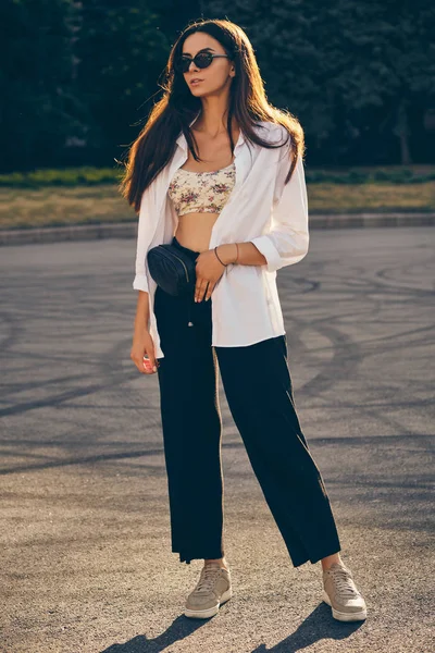 Retrato de uma menina de óculos escuros posando na cidade. Vestido em cima com estampa floral, camisa branca, calças pretas, bolsa de cintura e tênis . — Fotografia de Stock