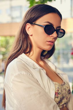 Yazlık terasın yakınlarında şehirde poz veren koyu güneş gözlüklü bir kızın portresi. Üstünde çiçek deseni, beyaz gömlek, siyah pantolon, bel çantası vardı..